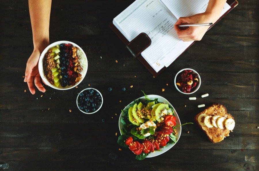 Desayuno saludable con bol de açaí, ensalada de frutas, tostada y suplementos junto a un diario de comidas